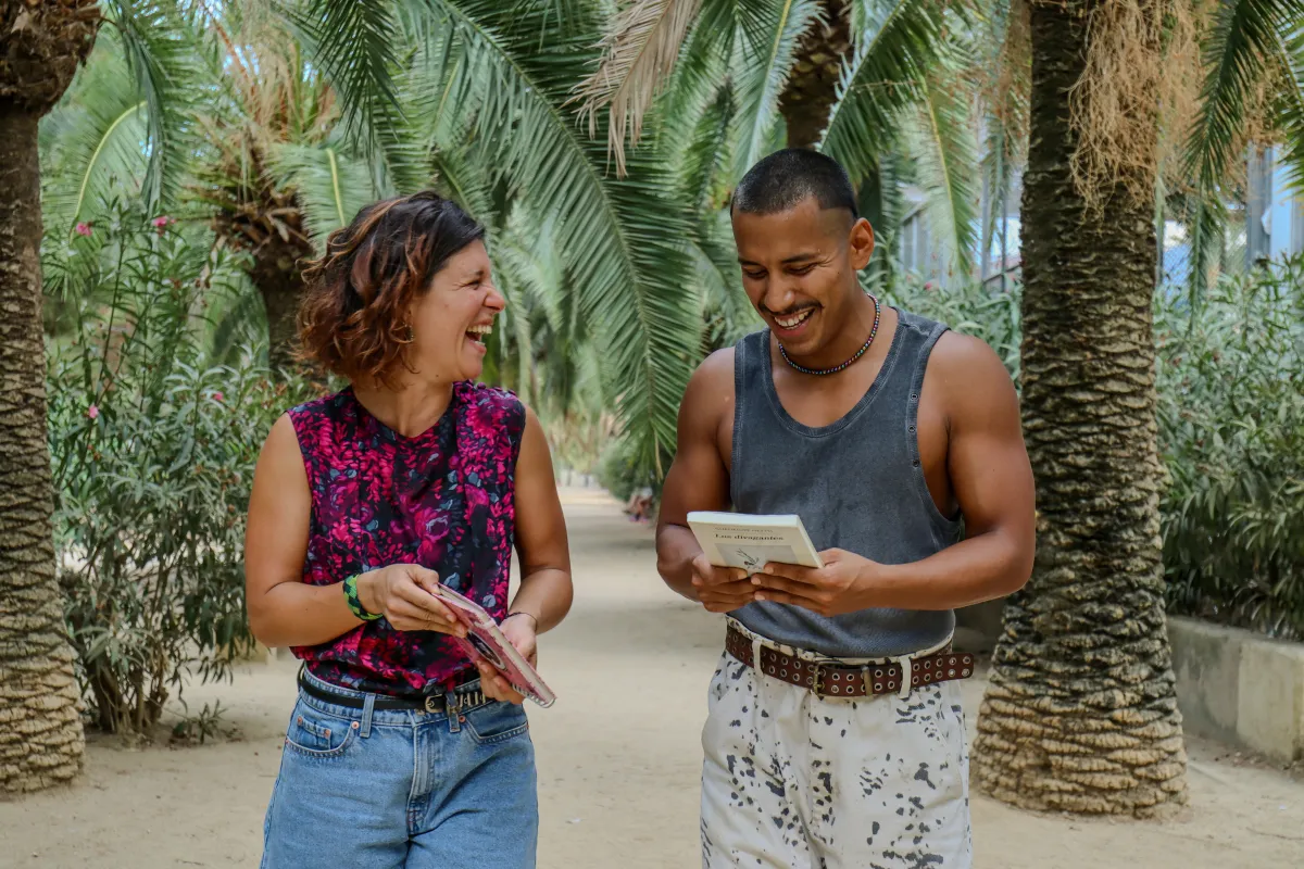 Emi and Nano walking under palm trees in a Barcelona park, laughing over a book